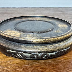May include: A dark brown wooden pedestal with a carved design around the edge. The pedestal has a slightly raised lip around the top.