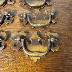 May include: A collection of antique-style brass drawer pulls featuring a curved, swinging handle attached to a decorative, ornate backplate with scrolled edges and a weathered, darkened patina finish, arranged on a wooden surface.