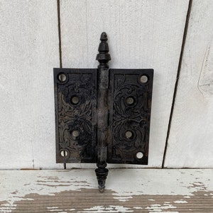 May include: Black cast iron door hinge with ornate floral design. The hinge has a pointed top and is attached to a white wooden surface.