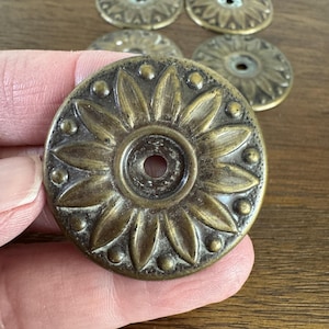 May include: A set of eight antique brass decorative accents with a floral design. Each accent features a raised center circle with a small hole in the center. The accents have a worn, aged look.