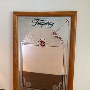 May include: A framed mirror with the Tanqueray logo in black script at the top. The mirror has a decorative etched design and a rectangular space for text. The frame is made of light brown wood.