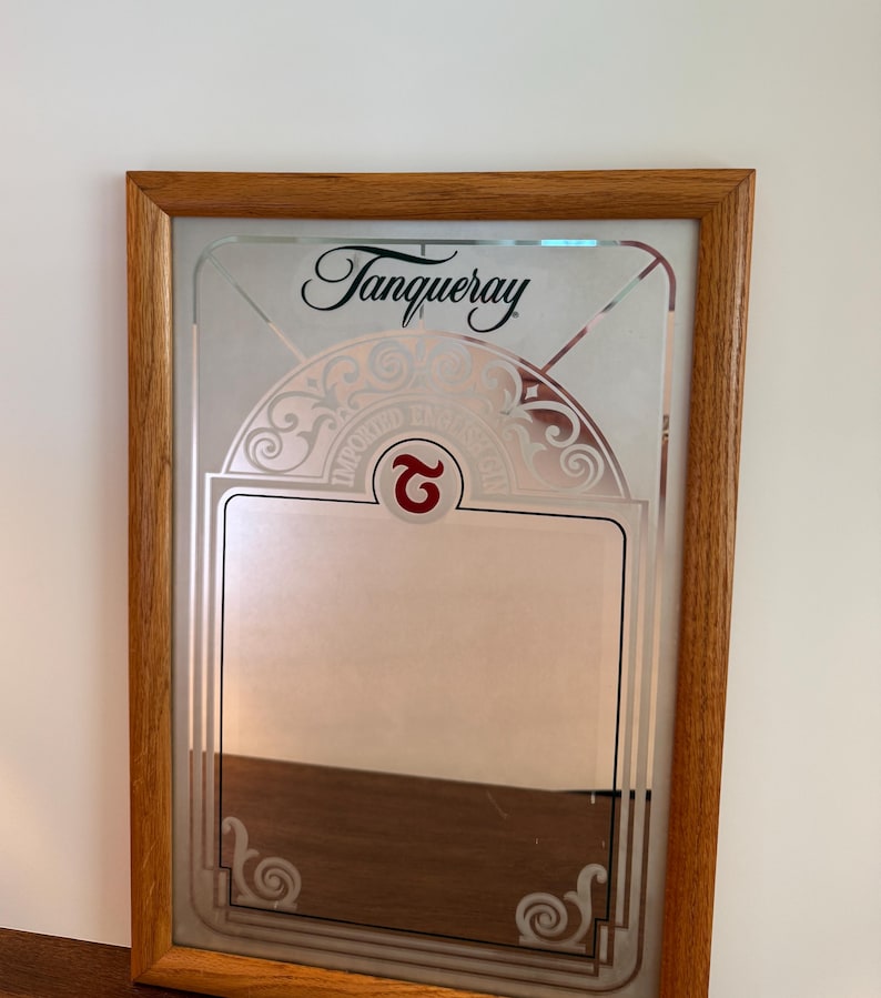 May include: A framed mirror with the Tanqueray logo in black script at the top. The mirror has a decorative etched design and a rectangular space for text. The frame is made of light brown wood.