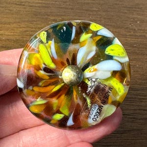 May include: A round glass coaster featuring a multi-colored pattern of yellow, white, and amber glass shards. A small metallic center stud is present. A clear sticker on the surface reads: MADE IN MEXICO FABRIQUE AU MEXIQUE HAND WASH ONLY.