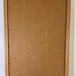 May include: Back of a framed picture with a light brown wooden frame. The backing is cardboard. The frame has a metal sawtooth hanger. There is a label with text in the bottom left corner and another label in the bottom right corner.