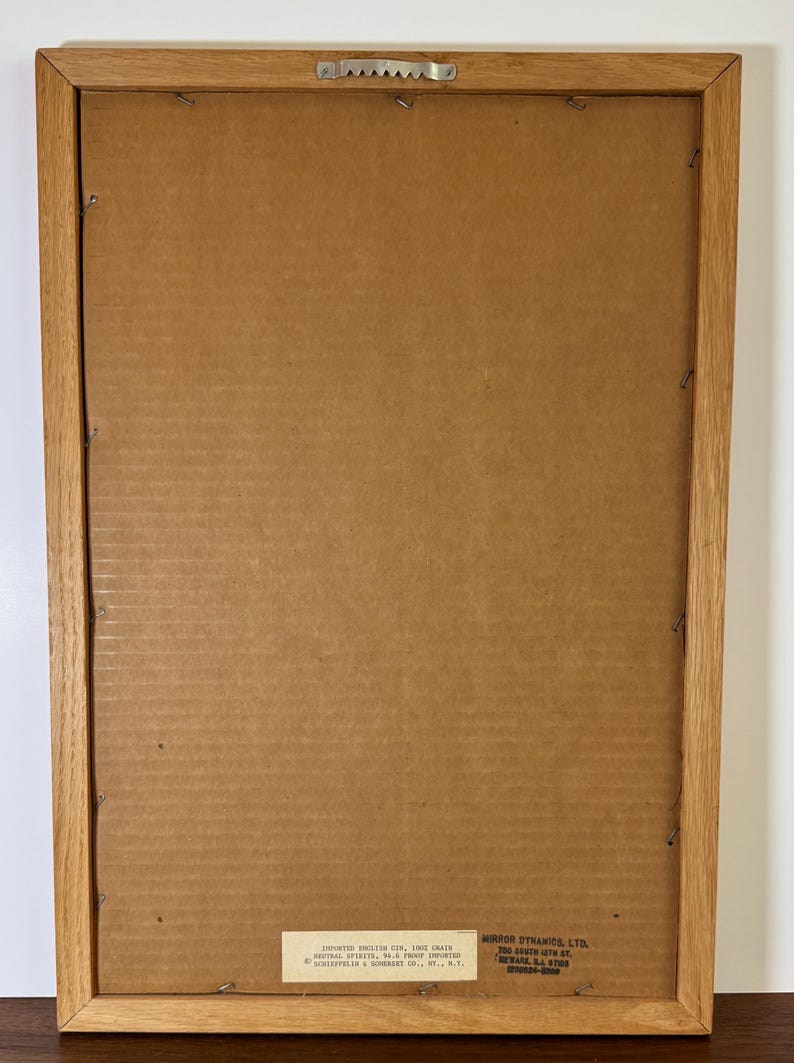 May include: Back of a framed picture with a light brown wooden frame. The backing is cardboard. The frame has a metal sawtooth hanger. There is a label with text in the bottom left corner and another label in the bottom right corner.