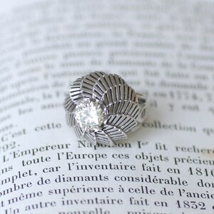 Puede incluir: Un anillo de plata con un gran diamante transparente engastado en un diseño con forma de hoja. El anillo está sobre una página de un libro con texto sobre diamantes.