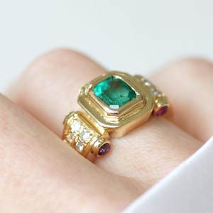Può includere: Un anello d'oro con un grande smeraldo quadrato come pietra centrale. L'anello è impreziosito da piccoli diamanti e due pietre rosse. L'anello è indossato su un dito, con tessuto bianco sullo sfondo.