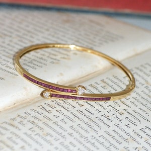 Può includere: Un braccialetto rigido in oro con una fila di piccole pietre preziose rosse e due accenti a forma di cuore. Il braccialetto è appoggiato su una pagina di un libro.