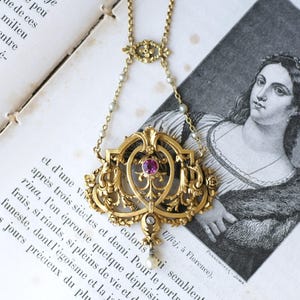 Peut inclure: Un collier pendentif en or avec un design ajouré complexe, une pierre précieuse rose ronde au centre, un petit diamant et une perle pendante. La chaîne est ornée de minuscules perles de semence et d'un connecteur décoratif en or.