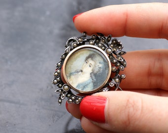 May include: A silver and gold brooch with a small portrait of a woman in a white dress. The brooch is decorated with small pearls and a floral design.