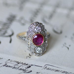 Könnte beinhalten: Ein Vintage-Ring mit einem großen, runden, roten Rubin, der von kleineren Diamanten umgeben ist. Der Ring ist in Silber und Gold gefasst.