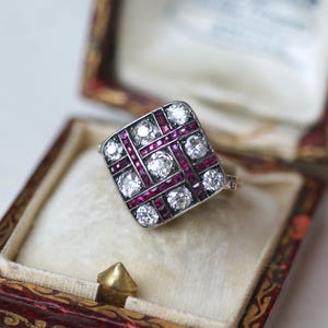 May include: A square-shaped ring with a geometric design featuring a grid of small diamonds and ruby accents. The ring is set in silver and is displayed in a red velvet ring box.