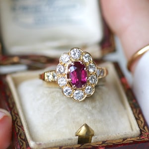 Peut inclure: Une bague en or avec un gros rubis ovale entouré d'un halo de petits diamants. La bague est dans un écrin à bijoux rouge et or.