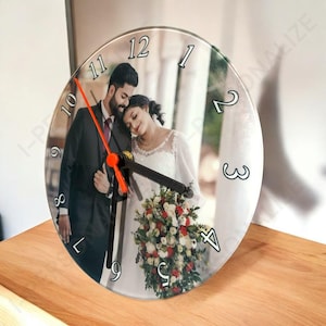 May include: A circular, clear glass clock featuring a photograph of a couple in formal wear. The clock has black numerals and black and orange hands. It rests on a wooden surface.