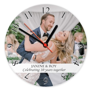 May include: A circular wall clock with a photograph of a couple. The clock face has black numerals, black and red hands, and the text "JANINE & ROY Celebrating 10 years together."