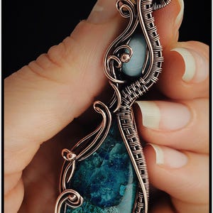May include: Copper wire-wrapped pendant with a teardrop-shaped, teal-colored stone. The pendant has a detailed, woven wire design and a smaller, round, light blue stone at the top. The pendant is held in a hand against a black background. The text "KELLY J JEWELLERY" is at the bottom.