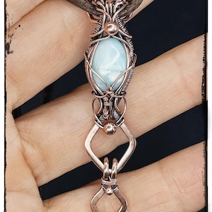 May include: A handcrafted copper pendant with a light blue oval gemstone, intricately wire-wrapped. It hangs from a brown cord. The text "KELLY J JEWELLERY" is at the bottom.