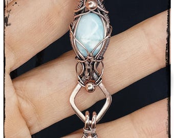 May include: A handcrafted copper pendant featuring a light blue oval gemstone, intricately wire-wrapped. The pendant hangs from a brown cord. The text "KELLY J JEWELLERY" is at the bottom.