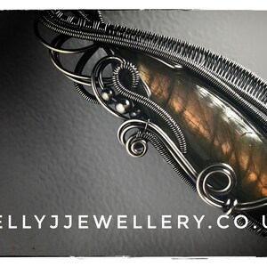 K&ouml;nnte beinhalten: Ein silberner, drahtgewickelter Anh&auml;nger mit einem braunen und goldenen Labradorit-Edelstein. Der Anh&auml;nger ist vor einem dunkelgrauen Hintergrund platziert. Der Text "KELLYJJWELLERY.CO.UK" ist am unteren Rand des Bildes sichtbar.