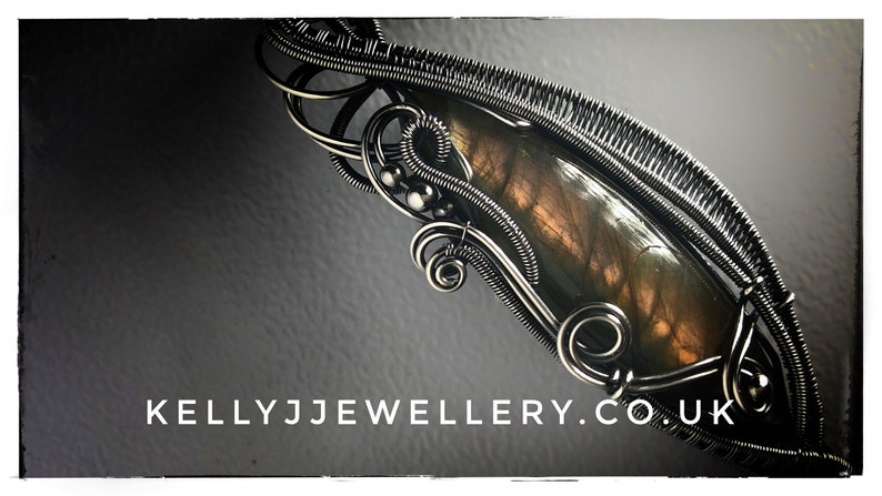 K&ouml;nnte beinhalten: Ein silberner, drahtgewickelter Anh&auml;nger mit einem braunen und goldenen Labradorit-Edelstein. Der Anh&auml;nger ist vor einem dunkelgrauen Hintergrund platziert. Der Text "KELLYJJWELLERY.CO.UK" ist am unteren Rand des Bildes sichtbar.