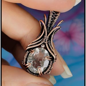 May include: A pendant featuring an oval-shaped clear gemstone with dark inclusions, encased in intricate copper wire wrapping. The metalwork displays a dark, oxidised finish with bright copper highlights. The text KELLY J JEWELLERY is visible.