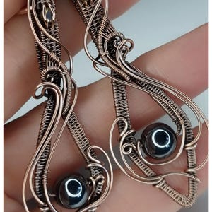 May include: A pair of copper wire earrings with a black hematite bead in each. The earrings are in the shape of a teardrop with intricate wirework.