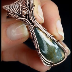 May include: A handcrafted copper wire pendant with a teardrop-shaped green and blue stone. The wirework forms an intricate cage around the stone. The text "KELLY J JEWELLERY" is at the bottom.