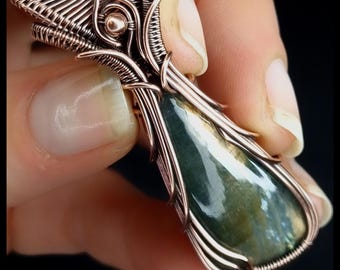 May include: A handcrafted copper wire pendant with a teardrop-shaped green and blue stone. The wirework forms an intricate cage around the stone. The text "KELLY J JEWELLERY" is at the bottom.