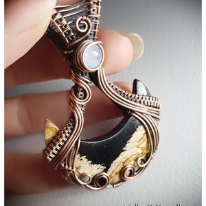 K&ouml;nnte beinhalten: Ein mit Kupferdraht umwickelter Anh&auml;nger mit einem schwarzen und wei&szlig;en Achat-Stein und einem Mondstein-Akzent. Der Anh&auml;nger ist handgefertigt und hat ein einzigartiges, filigranes Design.