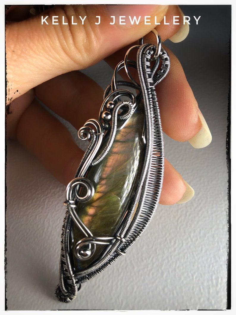 K&ouml;nnte beinhalten: Ein silberner, drahtgewickelter Anh&auml;nger mit einem gr&uuml;nen und irisierenden Labradorit-Edelstein. Der Anh&auml;nger hat die Form einer Tr&auml;ne und ein zartes, filigranes Design.