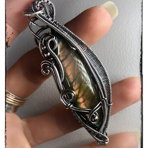 K&ouml;nnte beinhalten: Ein Anh&auml;nger aus silbernem Draht, der einen gr&uuml;n-goldenen Labradorit-Edelstein umh&uuml;llt. Der Anh&auml;nger wird in einer Hand gehalten.