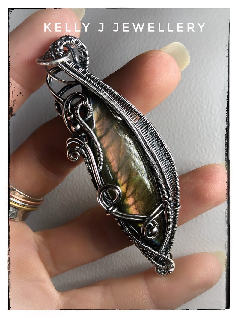 K&ouml;nnte beinhalten: Ein Anh&auml;nger aus silbernem Draht, der einen gr&uuml;n-goldenen Labradorit-Edelstein umh&uuml;llt. Der Anh&auml;nger wird in einer Hand gehalten.