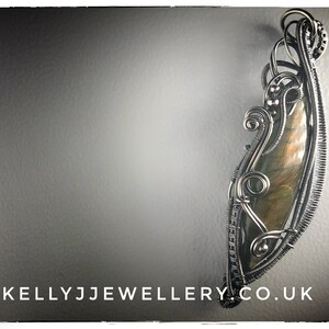 K&ouml;nnte beinhalten: Ein Anh&auml;nger aus silbernem Draht, der einen gro&szlig;en, schillernden Labradorit-Edelstein umh&uuml;llt. Der Draht ist zu einem komplizierten Muster gedreht und gekr&auml;uselt.