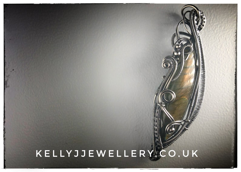 K&ouml;nnte beinhalten: Ein Anh&auml;nger aus silbernem Draht, der einen gro&szlig;en, schillernden Labradorit-Edelstein umh&uuml;llt. Der Draht ist zu einem komplizierten Muster gedreht und gekr&auml;uselt.