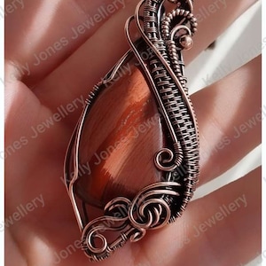 May include: A copper wire wrapped pendant featuring a red tiger's eye stone. The pendant is held in a hand with the text "KELLY JONES JEWELLERY" visible at the bottom of the image.