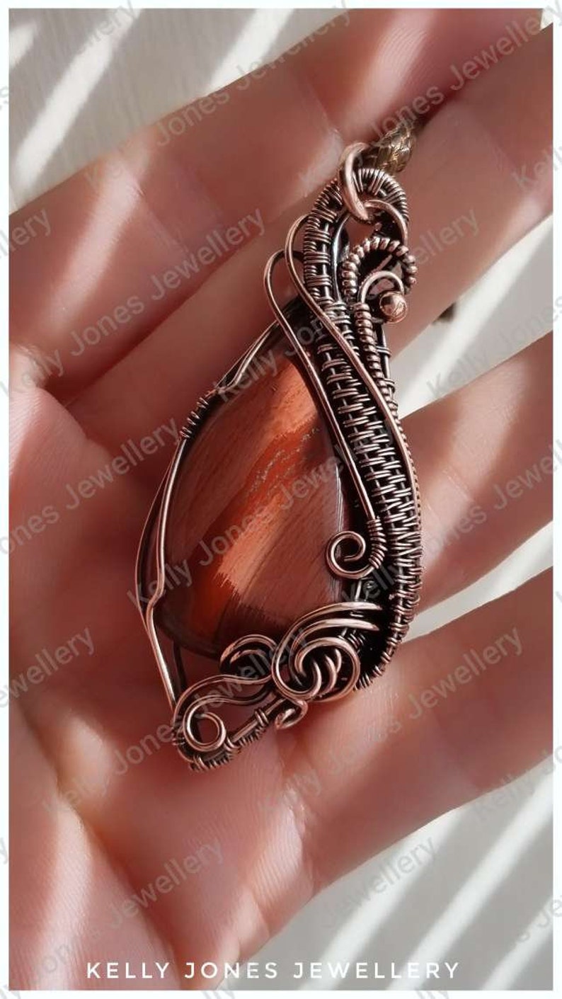 May include: A copper wire wrapped pendant featuring a red tiger's eye stone. The pendant is held in a hand with the text "KELLY JONES JEWELLERY" visible at the bottom of the image.