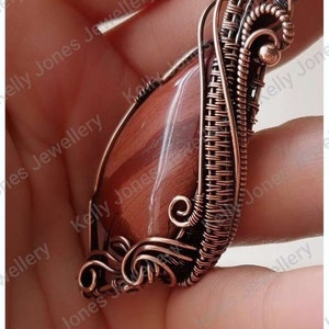 May include: A copper wire wrapped pendant featuring a red and brown gemstone. The pendant is held in a hand.