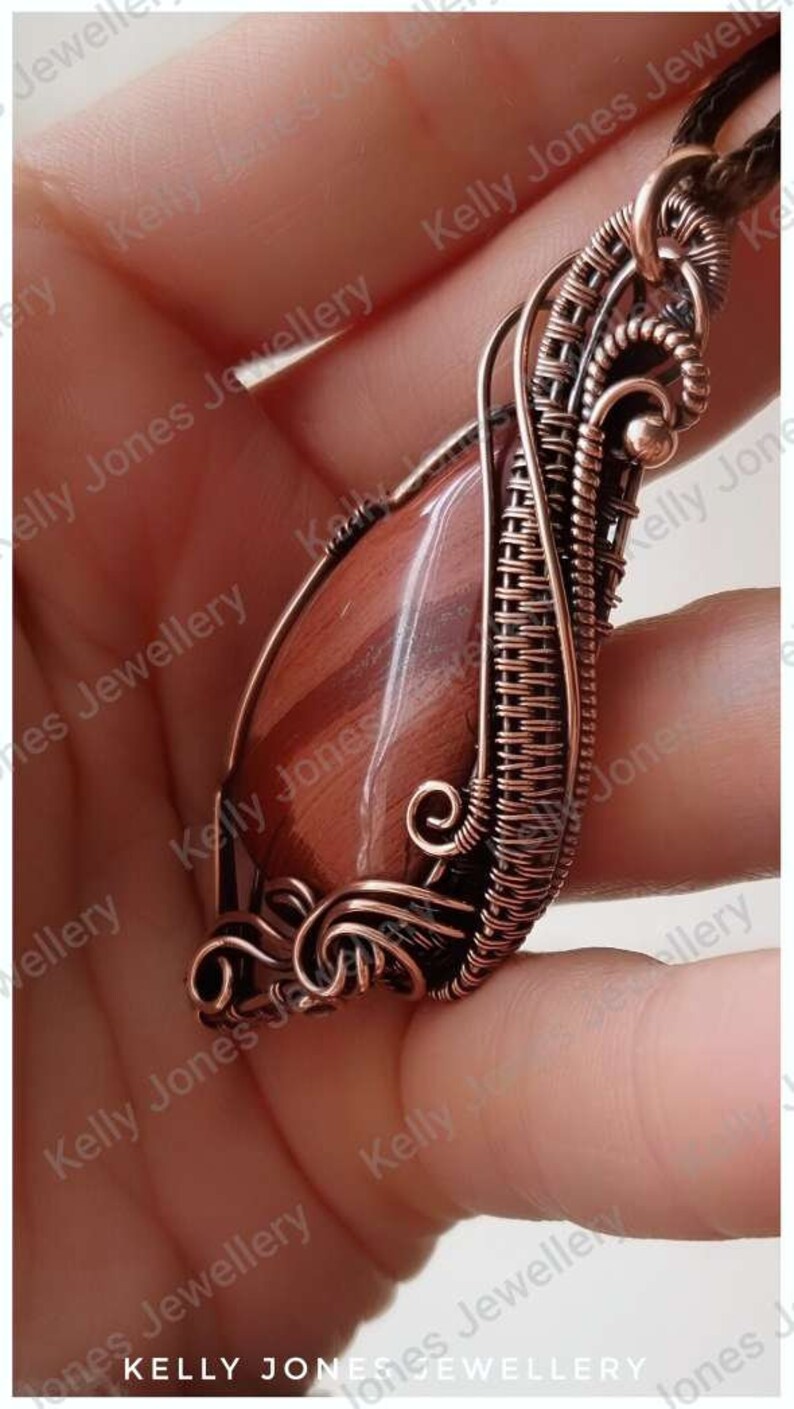 May include: A copper wire wrapped pendant featuring a red and brown gemstone. The pendant is held in a hand.
