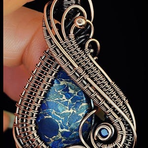 Könnte beinhalten: Handgefertigter Anhänger mit einem blauen Stein mit einzigartigem Muster, eingefasst in filigranem Kupferdraht. Die Drahtarbeit bildet ein künstlerisches Design, mit dem Text "KELLY J JEWELLERY" unten. Der Anhänger vor schwarzem Hintergrund.