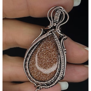 Könnte beinhalten: Ein handgefertigter Anhänger mit einem tropfenförmigen, funkelnden braunen Stein und einem weißen Halbmond-Design. Der Stein ist in aufwendiger kupferfarbener Drahtarbeit eingefasst. Der Anhänger wird von einer kleinen Schlaufe gehalten. Der Text "KELLY J JEWELLERY" steht unten.