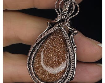 May include: A handcrafted pendant featuring a teardrop-shaped, sparkling brown stone with a white crescent design. The stone is encased in intricate copper-colored wirework. The pendant is held up by a small loop. The text "KELLY J JEWELLERY" is at the bottom.