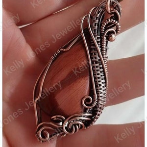 May include: A copper wire wrapped pendant featuring a red tiger's eye stone. The pendant is handmade and has a unique, intricate design.
