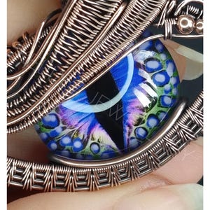 Op de afbeelding: Handgemaakte hanger met gedetailleerde koperdraadwikkeling. Het middelpunt is een glazen oog met blauwe, groene en paarse patronen. De hanger hangt aan een koperdraadketting. De tekst "KELLY J JEWELLERY" staat onderaan.