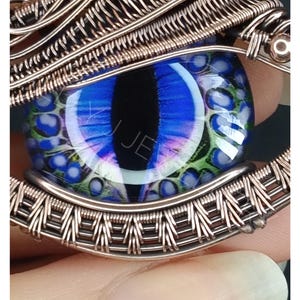 May include: A detailed close-up of a pendant with a blue and green glass eye cabochon, encased in copper wire. The eye has a black pupil and is surrounded by intricate wirework. The pendant is a unique piece of jewellery.