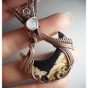 K&ouml;nnte beinhalten: Ein Anh&auml;nger aus Kupferdraht, der mit einem schwarzen und wei&szlig;en Achat-Stein mit einem Mondstein-Akzent umwickelt ist. Der Anh&auml;nger ist handgefertigt und hat ein einzigartiges, organisches Design.