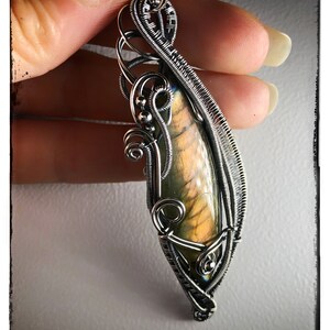 K&ouml;nnte beinhalten: Ein silberner, drahtgewickelter Anh&auml;nger mit einem perlmuttartigen Labradorit-Edelstein. Der Anh&auml;nger hat ein zartes, filigranes Design mit einem Wirbelmuster. Der Anh&auml;nger wird in einer Hand gehalten, und der Hintergrund ist eine verschwommene, graue Oberfl&auml;che. Der Text "KELLY J JEWELLERY" ist am unteren Rand des Bildes sichtbar.