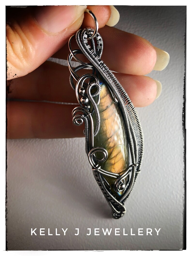 K&ouml;nnte beinhalten: Ein silberner, drahtgewickelter Anh&auml;nger mit einem perlmuttartigen Labradorit-Edelstein. Der Anh&auml;nger hat ein zartes, filigranes Design mit einem Wirbelmuster. Der Anh&auml;nger wird in einer Hand gehalten, und der Hintergrund ist eine verschwommene, graue Oberfl&auml;che. Der Text "KELLY J JEWELLERY" ist am unteren Rand des Bildes sichtbar.