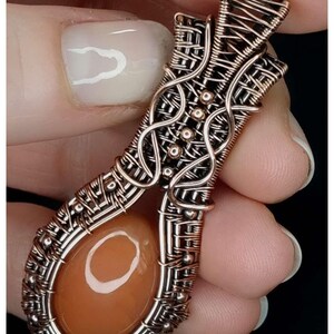 May include: A copper wire wrapped pendant with a peach-colored stone in the center. The pendant is made with a woven wire design and has small copper beads around the stone.