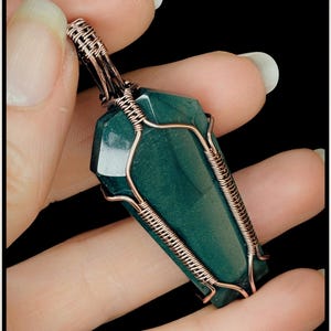 May include: A dark green gemstone pendant, intricately wrapped in copper wire. The gemstone has a rectangular, faceted shape. The wirework creates a detailed design around the stone. The pendant is held against a black backdrop, showcasing the jewellery.