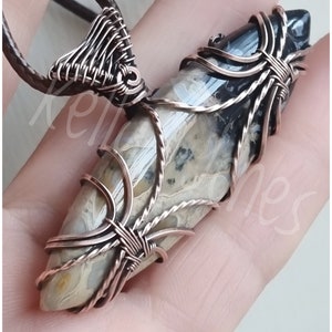 May include: A copper wire wrapped pendant featuring a light brown and black agate stone. The pendant is attached to a brown leather cord.